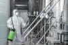 Factory Worker In Protective Suit With Spray Bags Disinfects Plant