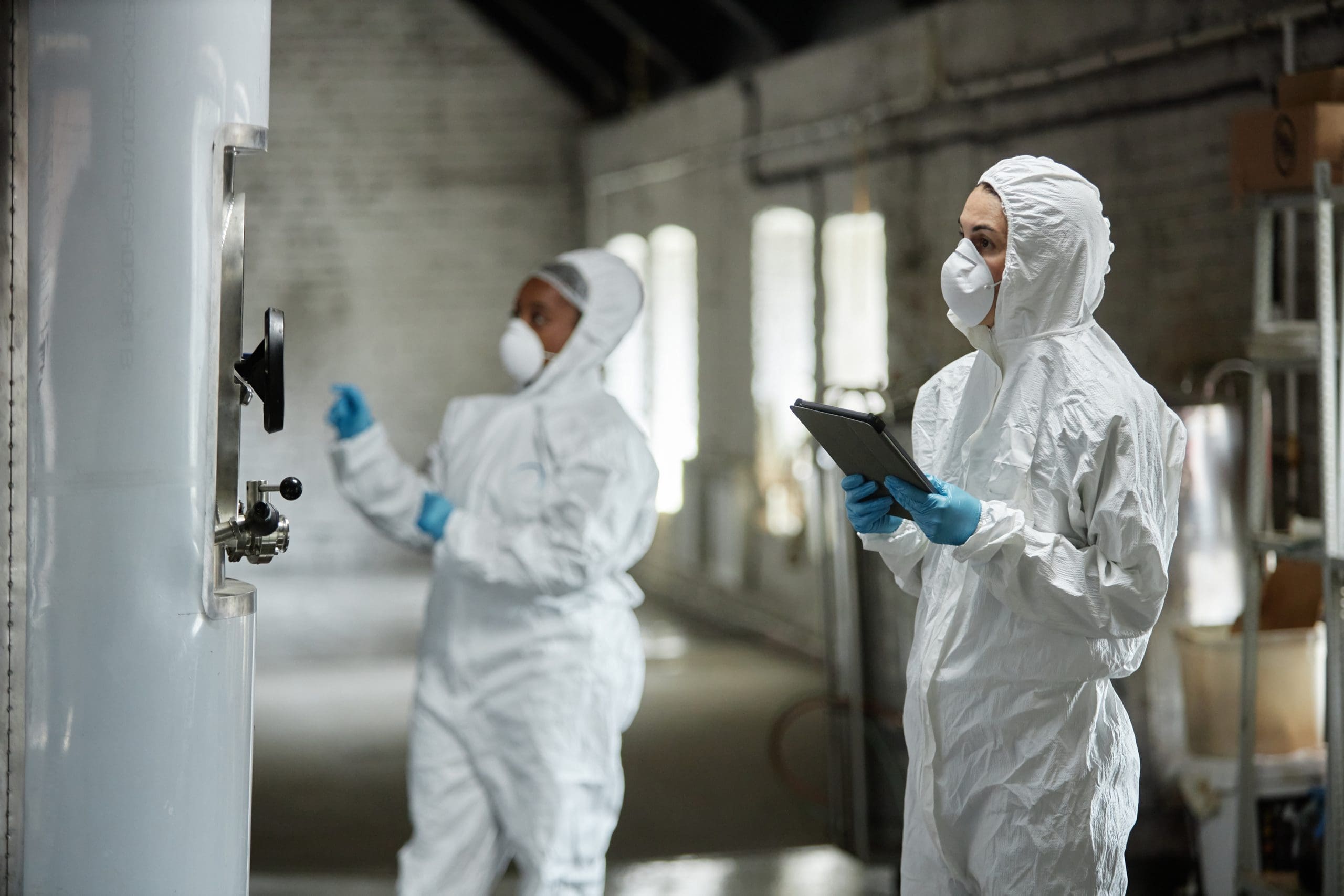 Two Female Supervisor In Hazmats Using Tablet Monitoring Fermentation In Tanks Indoors